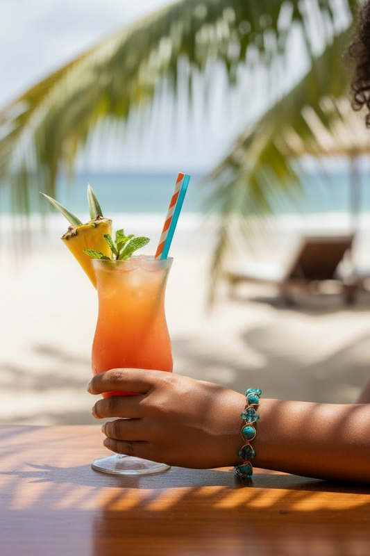 A person sits at a wooden table by the beach holding a tall tropical drink that fades from orange to pink, garnished with a pineapple wedge and mint leaves, with striped straws in the glass. On their wrist is a teal-colored bracelet featuring round teal stones alternating with faceted beads, secured in gold-toned wire. Palm trees and ocean water blur softly in the sunny background.