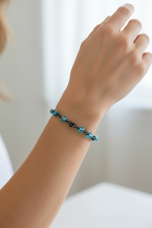 Close-up of a wrist wearing a bracelet made with black wire, featuring aqua rondelle beads arranged two per link on either side of a central love-knot detail. Each love-knot link is connected with double jump rings, creating a repeating, structured pattern around the wrist. Soft natural light highlights the aqua beads against a softly blurred neutral background.