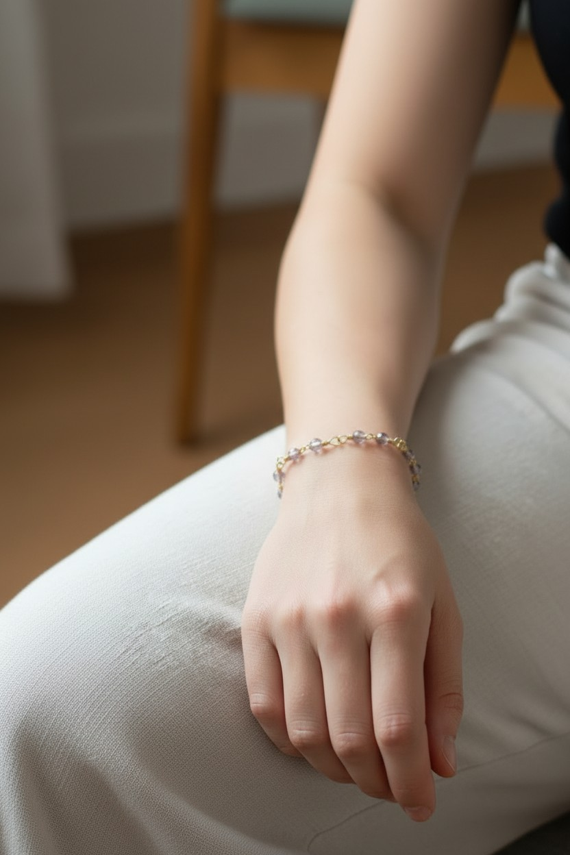 Close-up of a person’s hand resting on their knee while seated indoors. They wear a delicate bracelet made of small gray iridescent beads spaced along gold-colored wire, with subtle open square knot details between sections. Soft natural light highlights the beads’ shifting tones, and the background shows a blurred wooden chair and neutral interior.