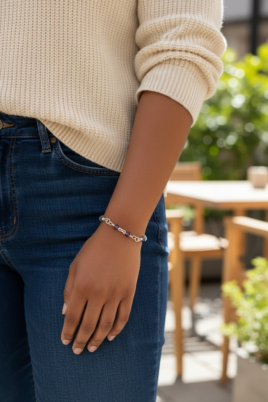 A person stands outdoors wearing a cream knit sweater and blue jeans, their arm relaxed at their side. On their wrist is a delicate bracelet made of silver-toned links, including open loose square-knot links, with small purple beads spaced on each side of the knot. Sunlight highlights the metal and beads while a wooden table and greenery blur in the background.