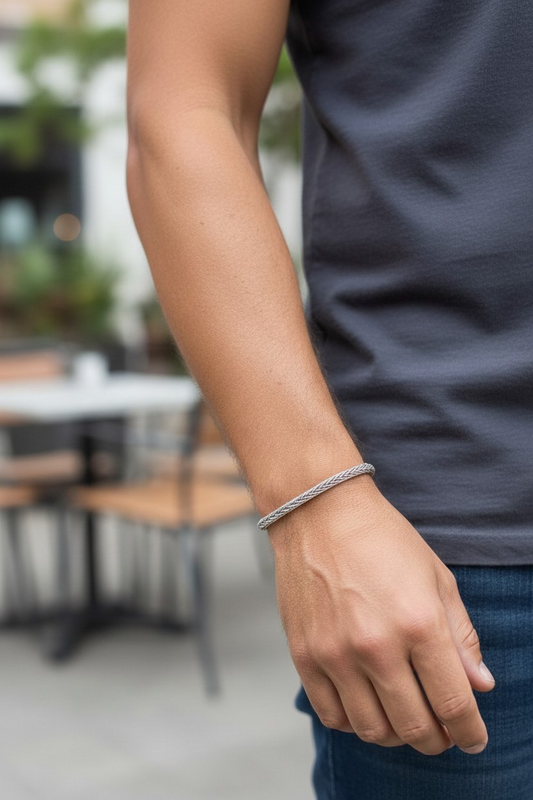 Person standing outdoors wearing a simple silver Trichinopoly weave bracelet on their wrist. The bracelet is a thin, tightly braided metallic weave forming a continuous loop with no visible clasp. The person is wearing a dark gray shirt and blue jeans, with blurred outdoor seating and greenery in the background.