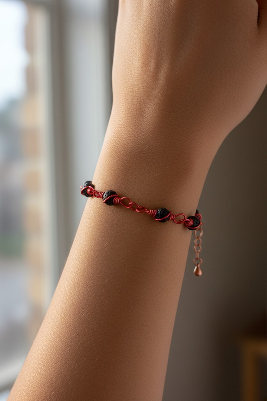 Close-up of a bracelet worn on a wrist near a window in soft natural light. The design features 6mm black lava beads spaced along the center, each secured by bright red wire twisted into a spiral pattern around the stones. The red wire continues into small linked sections and ends in a short extender chain with a small teardrop charm.