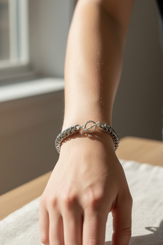 wrist wearing chainmail bracelet: tightly interlinked silver-toned rings forming a dense Full Persian 6-in-1 weave; toggle clasp resting at the top with twisted ring and straight bar; bracelet fitting close around the wrist; natural light from a nearby window casting soft highlights and shadows across the skin and metal; background softly blurred with a light surface and window frame visible