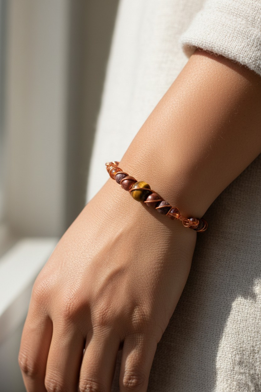 Close-up of a person’s hand and wrist in soft sunlight. The wrist wears a slim handcrafted bracelet made of twisted copper wire in a spiral wrap pattern, locking the beads into place. At the center is a bright copper-colored tiger’s eye stone, flanked by bronze-colored resin pearls; brown lava beads and dark amber colored rondelles continue along the bracelet.
