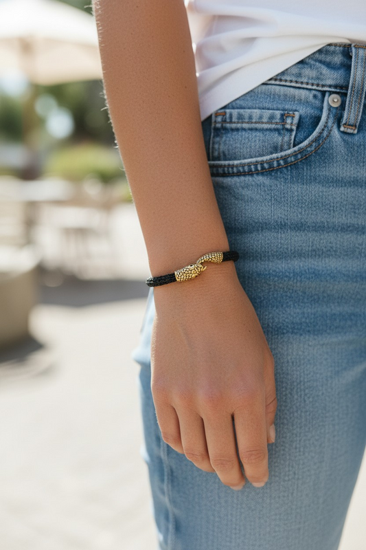 A Person standing outdoors wearing a black braided bracelet on their wrist. The bracelet features an Ouroboros-style gold-tone snake clasp at the center, where the snake’s tail hooks into the open snake head to form the closure. The person is wearing blue jeans and a white shirt, with the bracelet shown clearly against the wrist in natural daylight.