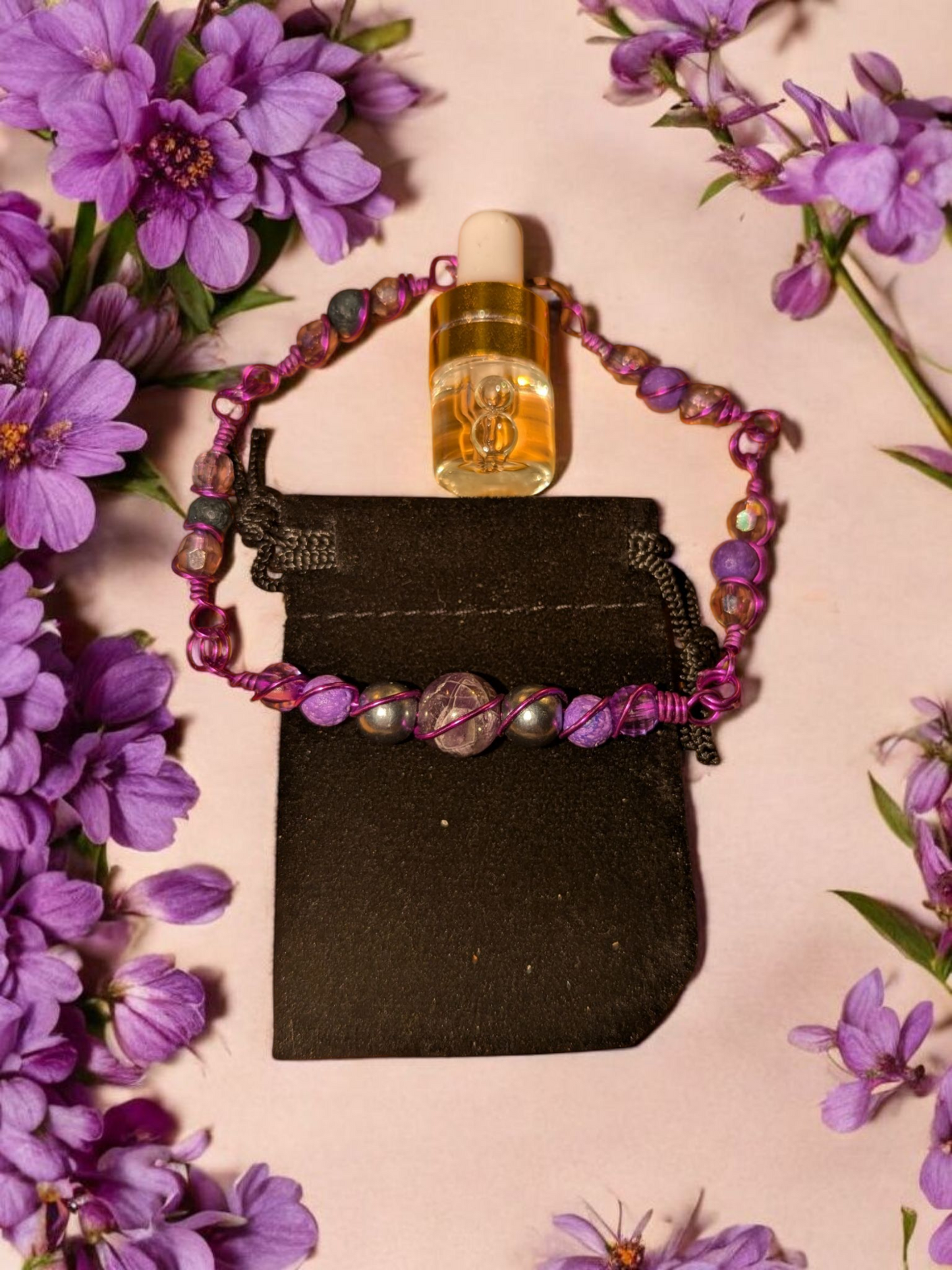 Handcrafted amethyst diffuser bracelet with dark resin pearls, purple lava stones, and purple hypoallergenic wire, displayed on a black velvet pouch surrounded by violet flowers.
