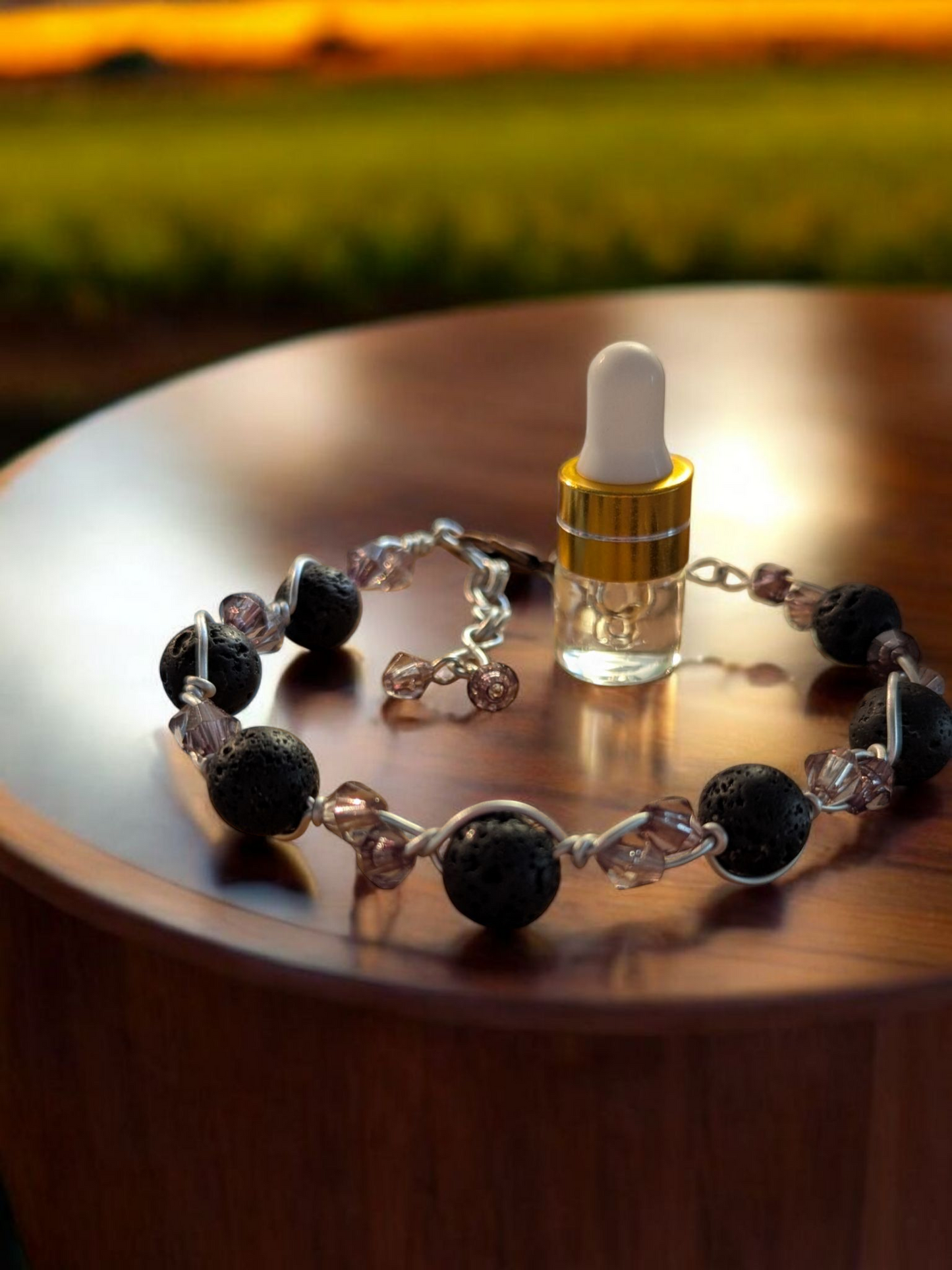 Handcrafted adjustable cuff diffuser bracelet with black lava stones and smokey gray resin bicone beads wrapped in silver-colored hypoallergenic wire, displayed on a polished wooden surface with an aromatherapy blend vial.
