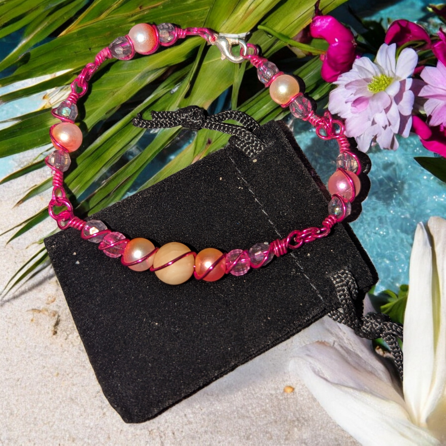 orbital wire-wrapped bracelet with warm jade and rose-toned beads and hot pink colored wire displayed on a black pouch near tropical flowers.