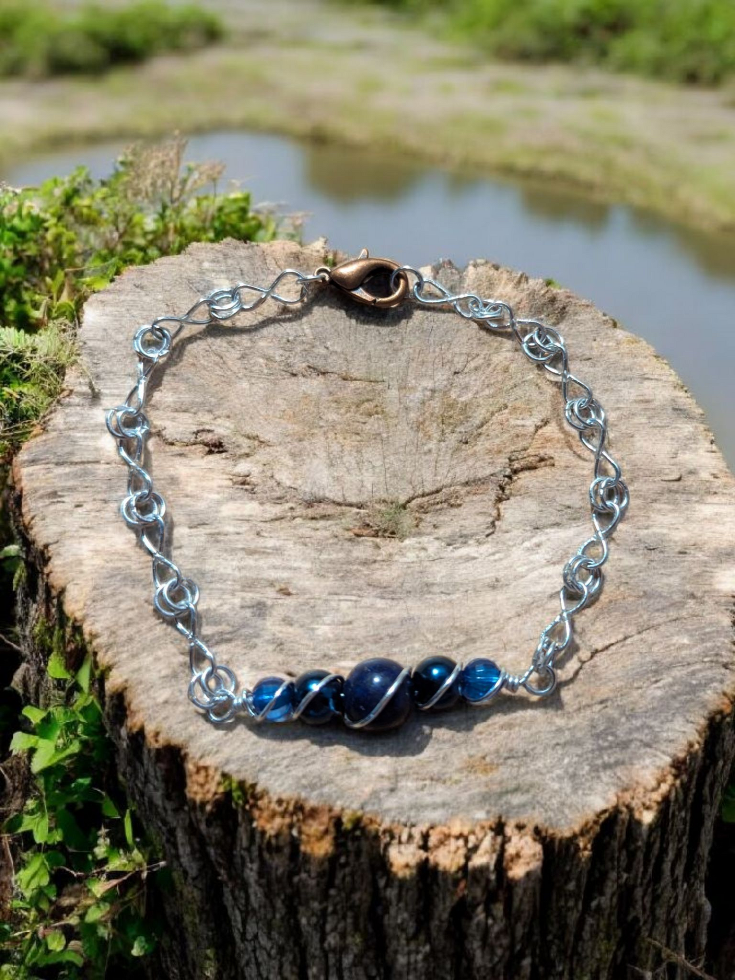 Handmade FireGryphonExclusive wire-wrapped link bracelet with deep blue beads and bronze clasp resting on a weathered tree stump, part of the Wireborne Series collection.