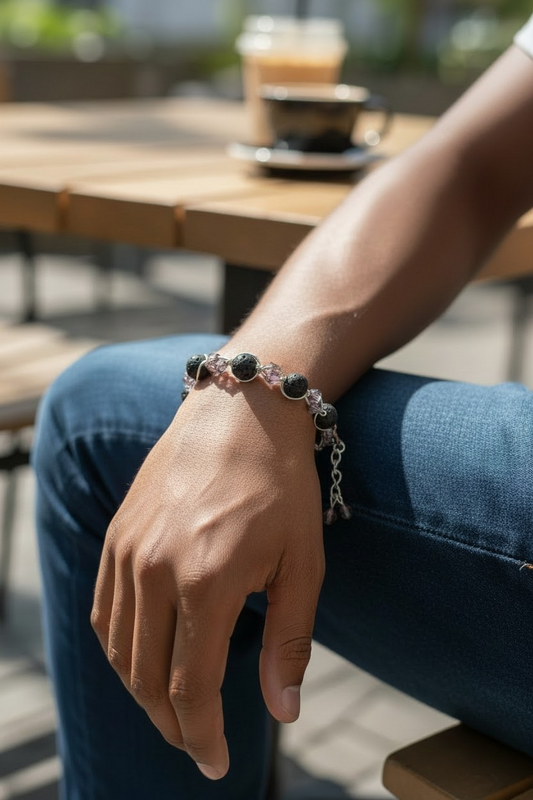 A male model sits outdoors at a wooden café table in bright sunlight, wearing a bracelet on his wrist. The bracelet features black lava stones alternating with faceted smokey gray bicone beads, connected by silver-toned wire and a short extension chain. He rests his hand on his knee while a blurred coffee cup and drink sit on the table in the background.