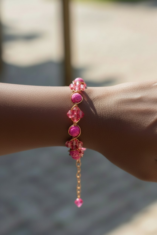 Close-up of a wrist wearing a bracelet made of bright pink lava stones alternating with faceted pink bicone beads. Each section is wrapped and secured in gold-toned wire, forming rounded links between the stones. A short gold extension chain with a small pink bead dangles from the clasp. The background is softly blurred in warm outdoor light.