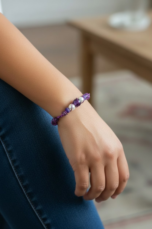 Close-up of a person’s forearm and hand resting over blue jeans. On the wrist is a slim bracelet made entirely of twisted wire in an orbital spiral pattern, locking beads into place. At the center is a small bead stack with a round translucent amethyst stone, flanked by two glossy white pearl-like beads, plus a smaller purple bead beside them. The wire ends are tightly wrapped to finish the bracelet.