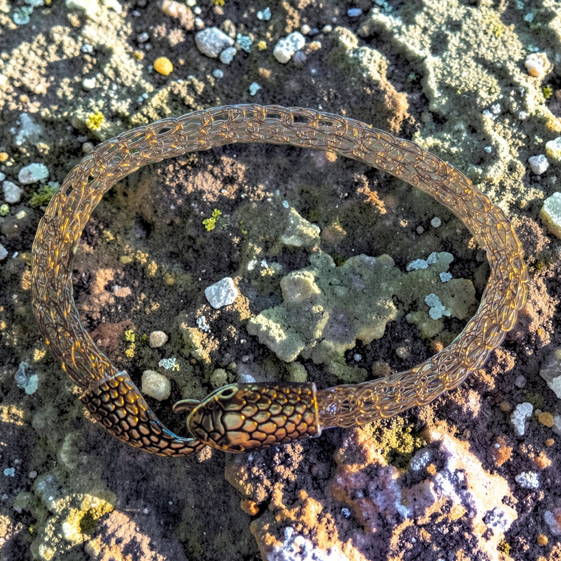 Close-up photo of a gold-toned Viking woven rope bracelet arranged in an oval on rough, gritty ground with scattered pebbles and pale green patches. At the front is a textured snake-head clasp, with the tail ending in a small hook that fastens by hooking into the snake head as if it’s eating the tail.