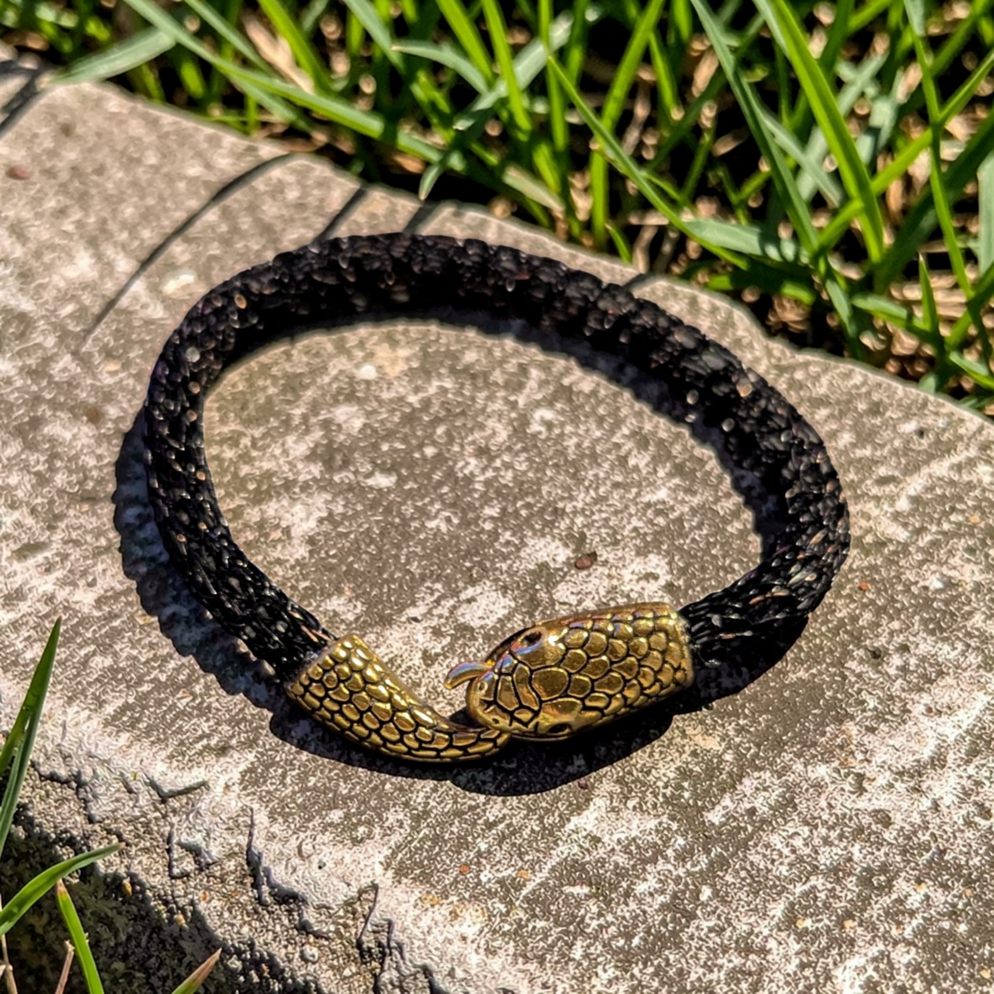 Black and copper braided bracelet with an Ouroboros-style gold-tone snake clasp resting on a textured stone surface outdoors. The snake tail hooks into the open snake head at the center to form the closure, with both pieces detailed in raised scale texture. Green grass borders the stone in the background.