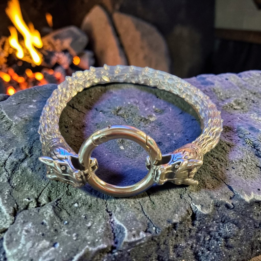 A woven bracelet made of 304 stainless steel rests on a rough stone surface beside a small fire. The bracelet features two detailed dragon heads facing each other, both biting the same round steel ring clasp at the center. Warm firelight glows in the background, highlighting the metallic weave and the sculpted details of the dragon heads.