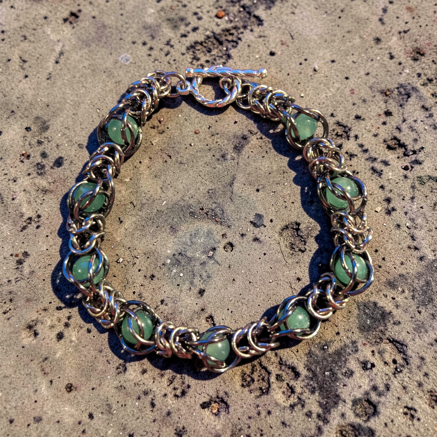 Erda, a circular chainmail bracelet laid flat on a pitted concrete surface; gunmetal black rings with a hematite-like finish forming a dense, fluid structure; soft green aventurine beads captured within the weave at intervals; each stone held in place by wrapped rings rather than strung; top shows a toggle clasp with bar and loop closure; overall look is structured metal with organic movement and evenly spaced green accents