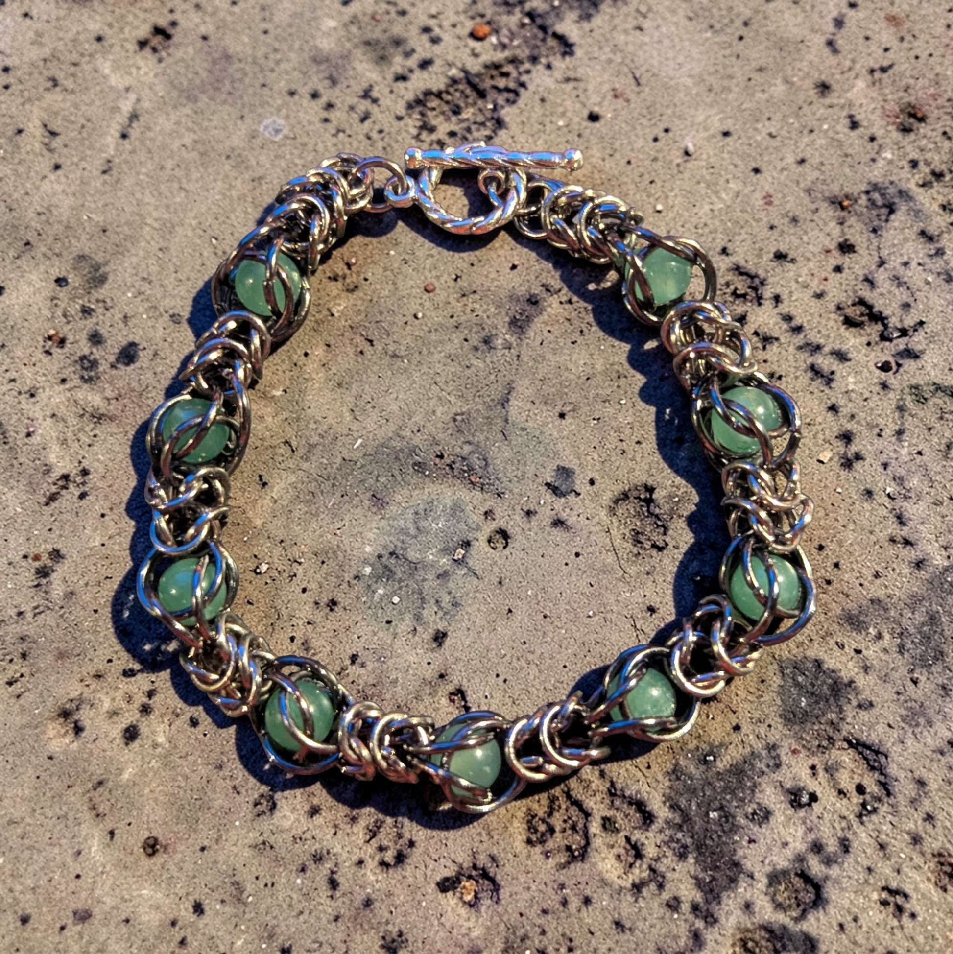 Erda, a circular chainmail bracelet laid flat on a pitted concrete surface; gunmetal black rings with a hematite-like finish forming a dense, fluid structure; soft green aventurine beads captured within the weave at intervals; each stone held in place by wrapped rings rather than strung; top shows a toggle clasp with bar and loop closure; overall look is structured metal with organic movement and evenly spaced green accents
