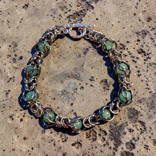 Erda, a circular chainmail bracelet laid flat on a pitted concrete surface; gunmetal black rings with a hematite-like finish forming a dense, fluid structure; soft green aventurine beads captured within the weave at intervals; each stone held in place by wrapped rings rather than strung; top shows a toggle clasp with bar and loop closure; overall look is structured metal with organic movement and evenly spaced green accents