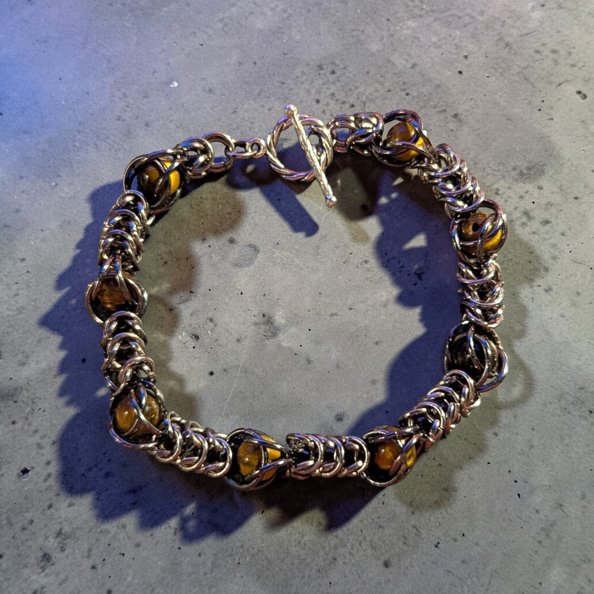 chainmail bracelet laid in a loose circle: tightly interlinked silver-toned rings forming a dense Full Persian style weave with Tiger’s Eye beads captured throughout the chainmail; warm brown and gold banding visible in the beads; toggle clasp at the top with twisted ring and straight bar; metal and beads catching light with soft reflections; set on a gray concrete surface with subtle texture and scattered speckling