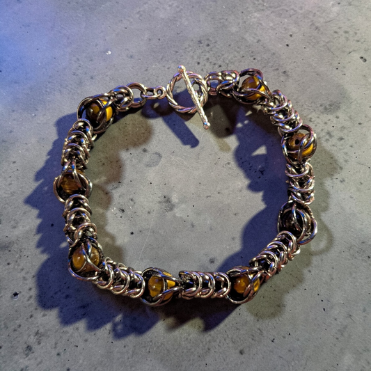 chainmail bracelet laid in a loose circle: tightly interlinked silver-toned rings forming a dense Full Persian style weave with Tiger’s Eye beads captured throughout the chainmail; warm brown and gold banding visible in the beads; toggle clasp at the top with twisted ring and straight bar; metal and beads catching light with soft reflections; set on a gray concrete surface with subtle texture and scattered speckling