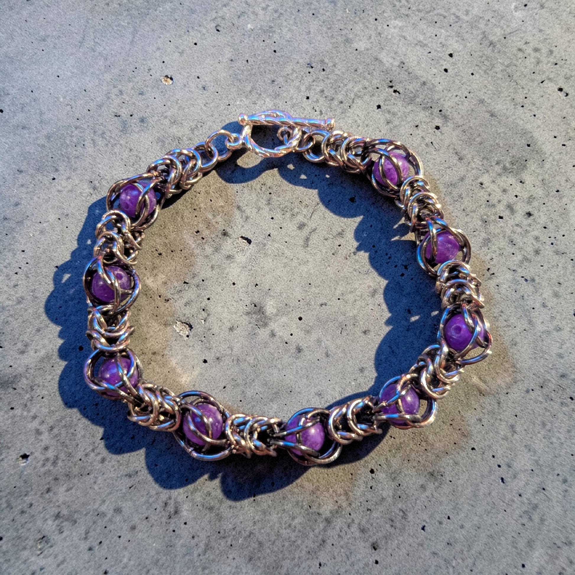 close view of a chainmail bracelet laid in a circle on a textured gray surface: silver full Persian 6-in-1 weave forming a fluid structure; purple amethyst beads captured within the rings at regular intervals; lobster clasp visible at the top; metal links reflect light with subtle highlights and shadowing