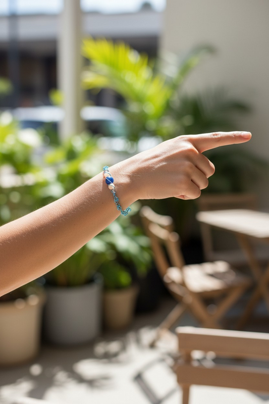 A close-up of a person’s outstretched arm pointing to the right in a bright indoor space filled with potted green plants and wooden café-style chairs. On the wrist is a blue and white beaded bracelet featuring a central deep blue round bead flanked by white beads and smaller aqua-toned beads, connected with delicate blue wire links. Sunlight streams through large windows, softly illuminating the bracelet and casting natural shadows.