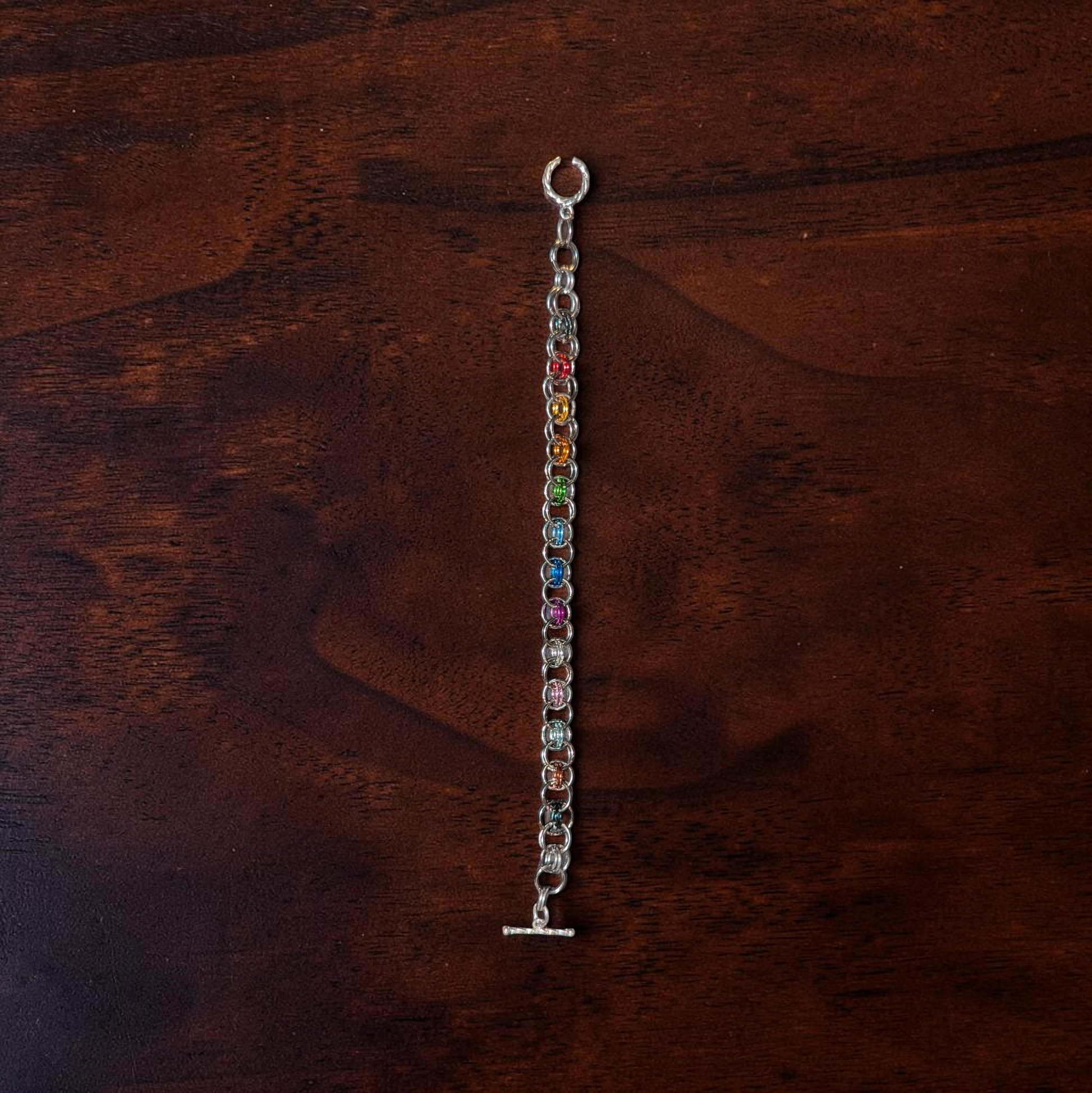 Close-up of Fyohl, a Helm Chain chainmail bracelet laid vertically on dark reddish-brown wood. The bracelet has larger hematite-like finished rings with a center color progression inspired by the Progress Pride flag: red, orange, yellow, green, blue, indigo, violet, white, pink, light blue, brown, and black. A round clasp is visible at the top and a toggle bar clasp at the bottom.