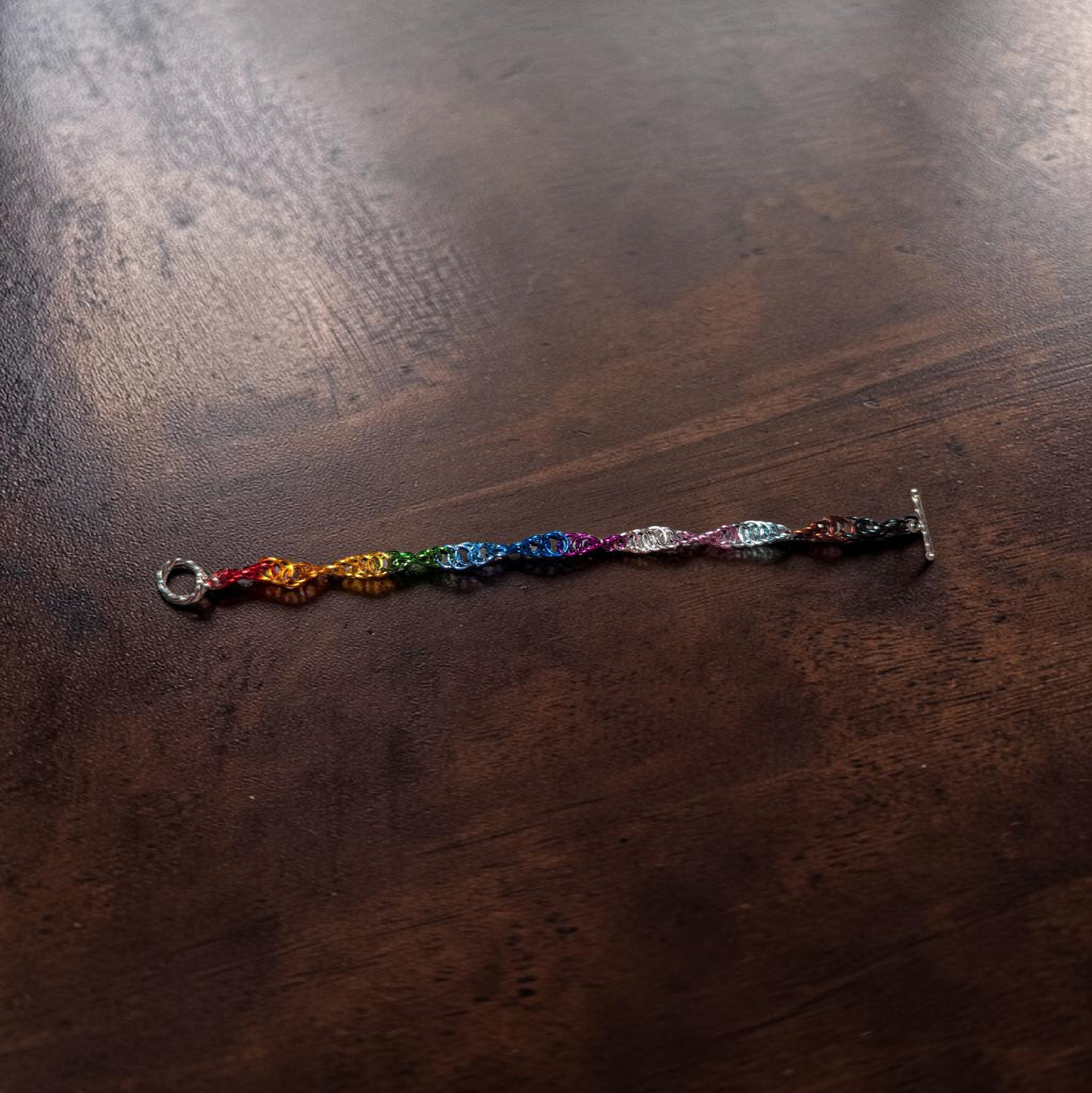 Close-up of Braut, a spiral 4-in-1 chainmail bracelet laid horizontally on dark reddish-brown wood. The bracelet uses a Progress Pride flag color progression: red, orange, yellow, green, blue, indigo, violet, white, light blue, pink, brown, and black. A round clasp is visible on one end and a toggle bar clasp on the other.