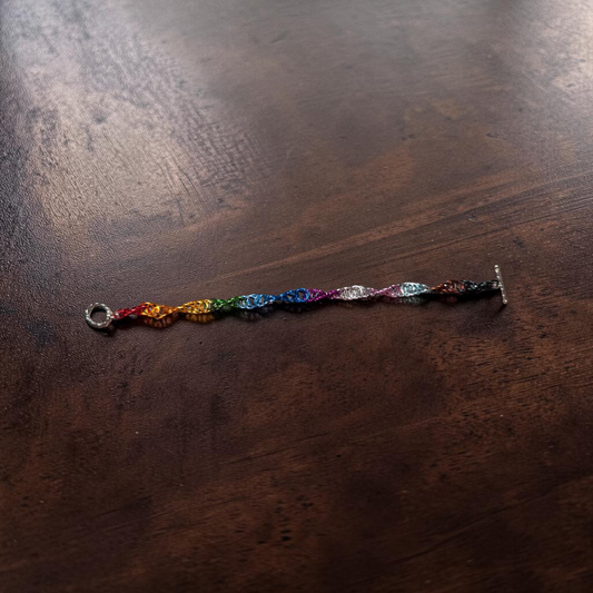 Close-up of Braut, a spiral 4-in-1 chainmail bracelet laid horizontally on dark reddish-brown wood. The bracelet uses a Progress Pride flag color progression: red, orange, yellow, green, blue, indigo, violet, white, light blue, pink, brown, and black. A round clasp is visible on one end and a toggle bar clasp on the other.