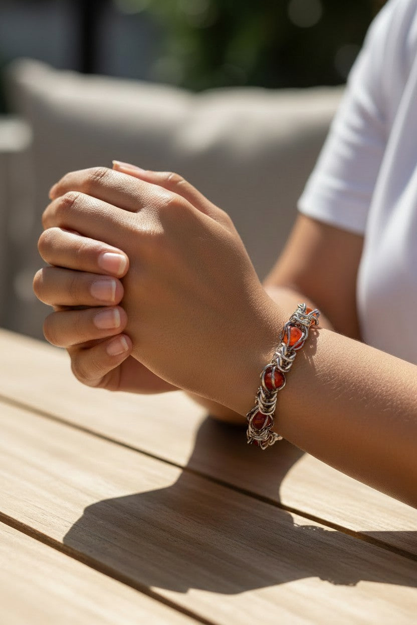 Virtual try-on photo of the Gun Metal Black chainmail bracelet on a medium skin-toned model’s wrist. The bracelet has a hematite-like reflective finish with glossy red agate beads trapped inside the chainmail pattern. The model’s hands are clasped on a sunlit wooden table, with a white shirt and softly blurred outdoor background.