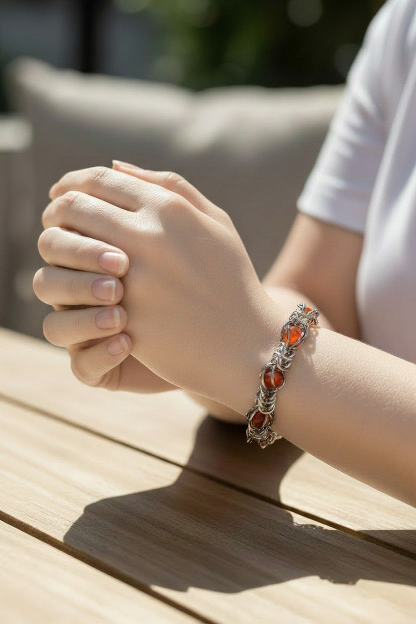 Virtual try-on photo of the Gun Metal Black chainmail bracelet on a pale skin-toned model’s wrist. The bracelet has a hematite-like reflective finish with glossy red agate beads trapped inside the chainmail pattern. The model’s hands are clasped on a sunlit wooden table, with a white shirt and softly blurred outdoor background.