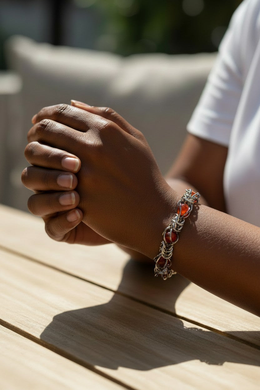 Virtual try-on photo of the Gun Metal Black chainmail bracelet on a dark skin-toned model’s wrist. The bracelet has a hematite-like reflective finish with glossy red agate beads trapped inside the chainmail pattern. The model’s hands are clasped on a sunlit wooden table, with a white shirt and softly blurred outdoor background.