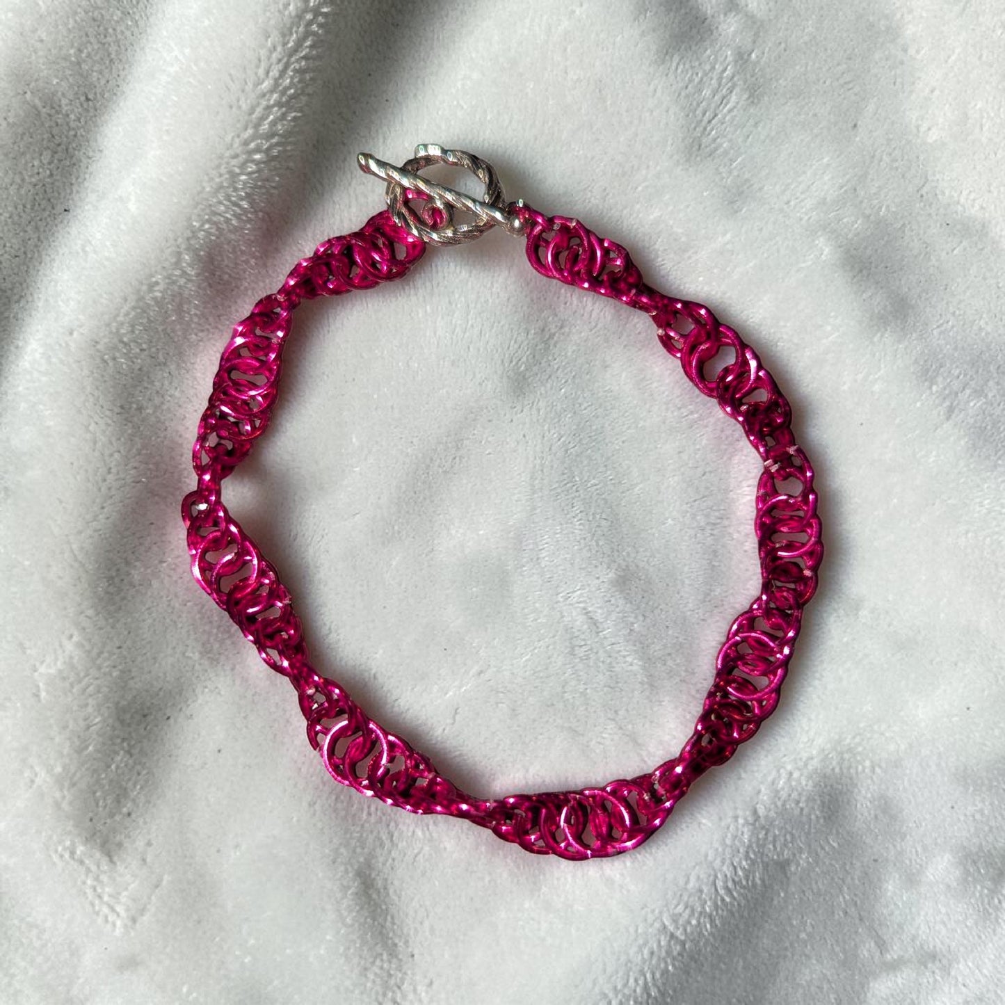 The Snerill spiral 4-in-1 chainmail bracelet in Fuchsia laid in a circle on white velvet, its vivid hot pink aluminum rings twisting in a continuous helix and finished with a silver toggle clasp at the top.