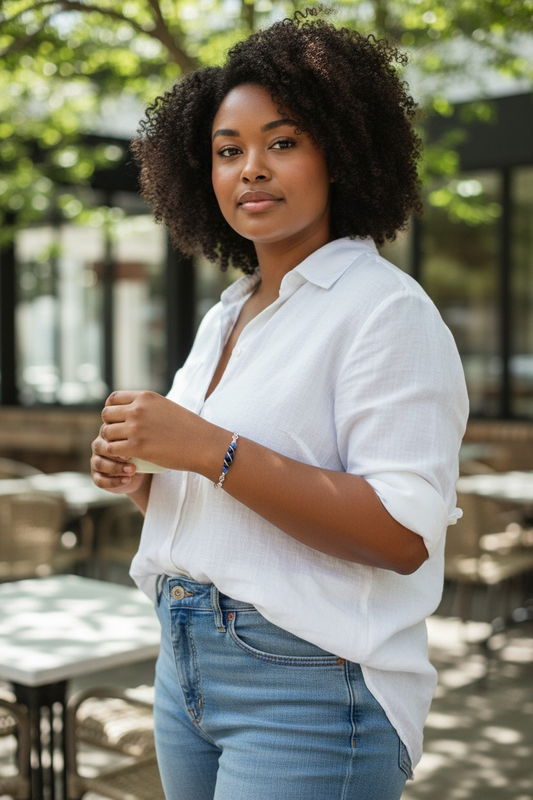 A woman with natural, shoulder-length curly hair stands outdoors at a café patio. She wears a loose white button-up blouse with rolled sleeves and light blue high-waisted jeans. She holds a small cup in both hands at chest level. On her right wrist is a blue beaded bracelet with silver accents. Behind her are metal café tables and chairs, green trees, and a modern glass-front building in soft daylight.