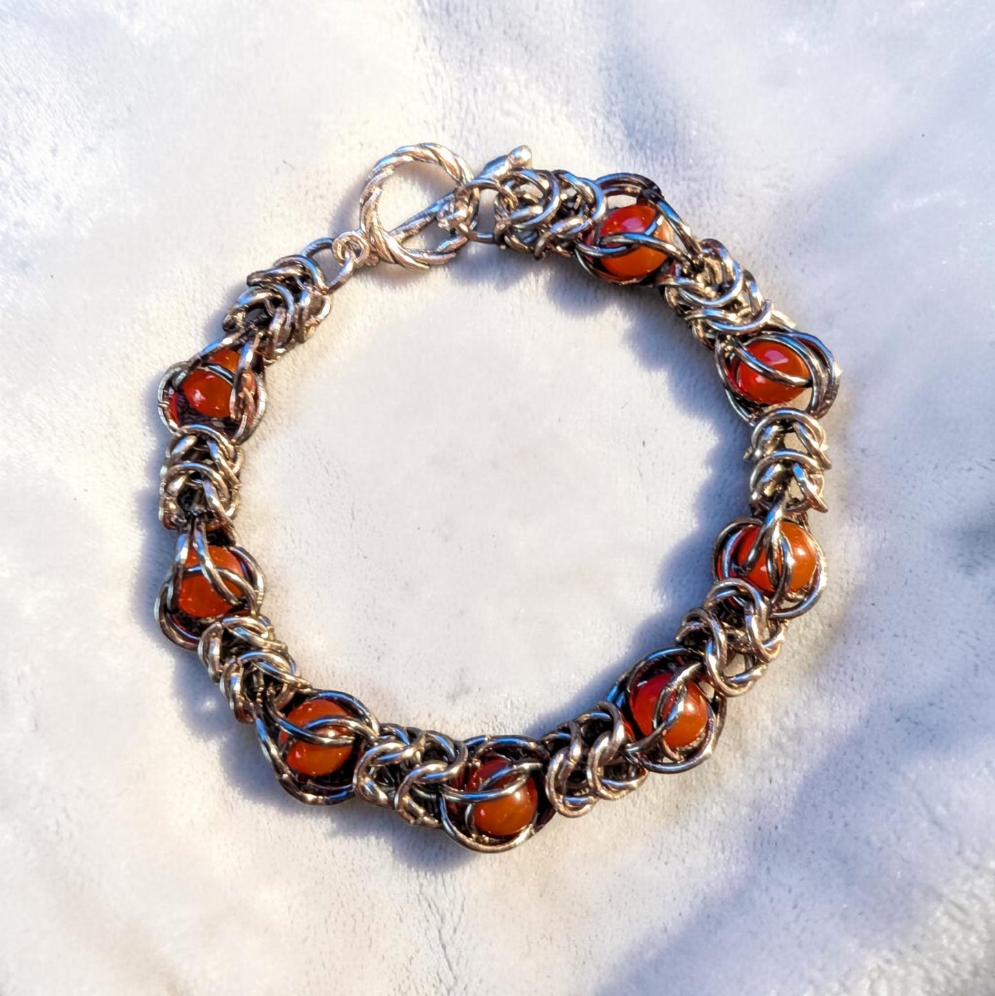 Gun Metal Black chainmail bracelet with a hematite-like reflective finish arranged in a circle on white velvet. Glossy red agate beads are trapped inside repeating sections of the chainmail pattern. A twisted toggle clasp sits at the top, and bright angled light creates sharp highlights and shadows across the bracelet.
