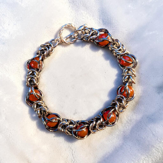 Gun Metal Black chainmail bracelet with a hematite-like reflective finish arranged in a circle on white velvet. Glossy red agate beads are trapped inside repeating sections of the chainmail pattern. A twisted toggle clasp sits at the top, and bright angled light creates sharp highlights and shadows across the bracelet.