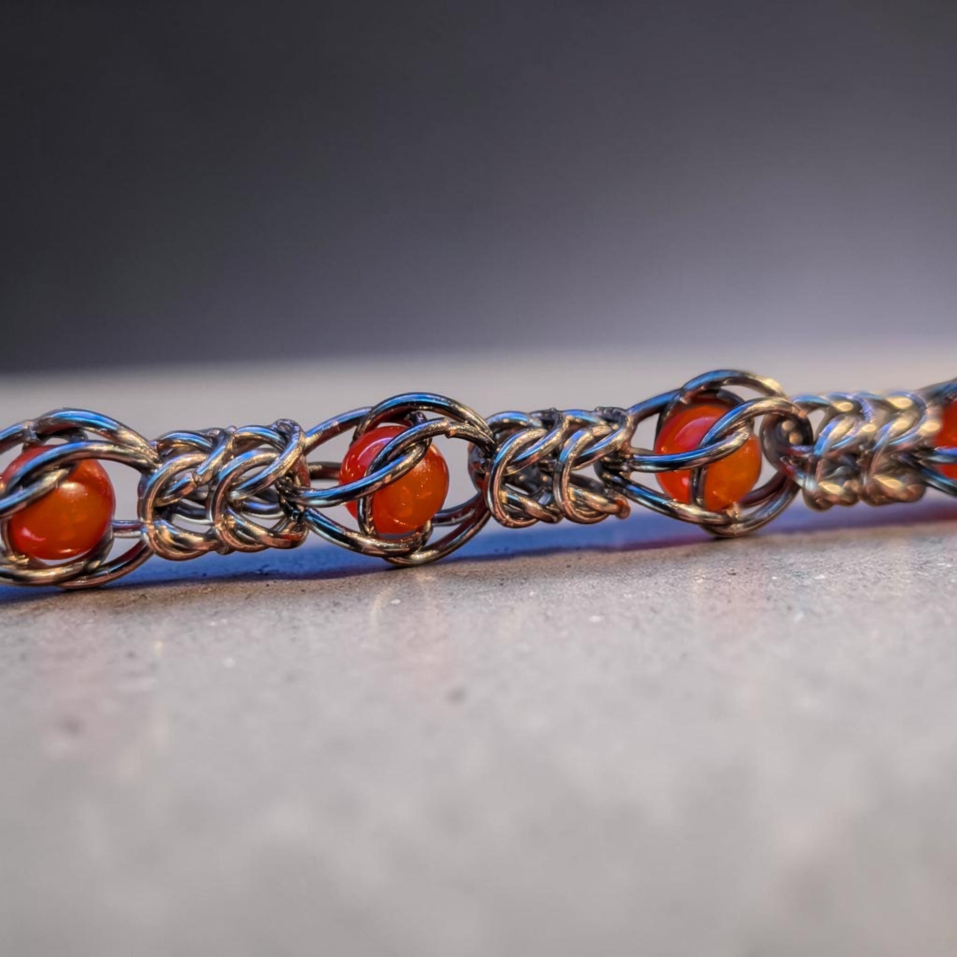 Macro close-up of a Gun Metal Black chainmail bracelet with a hematite-like reflective finish. Glossy red agate beads are trapped inside the repeating chainmail pattern, with the rings forming cage-like sections around each bead. The bracelet rests on a pale surface with shallow focus, bright metal highlights, and a softly blurred background.