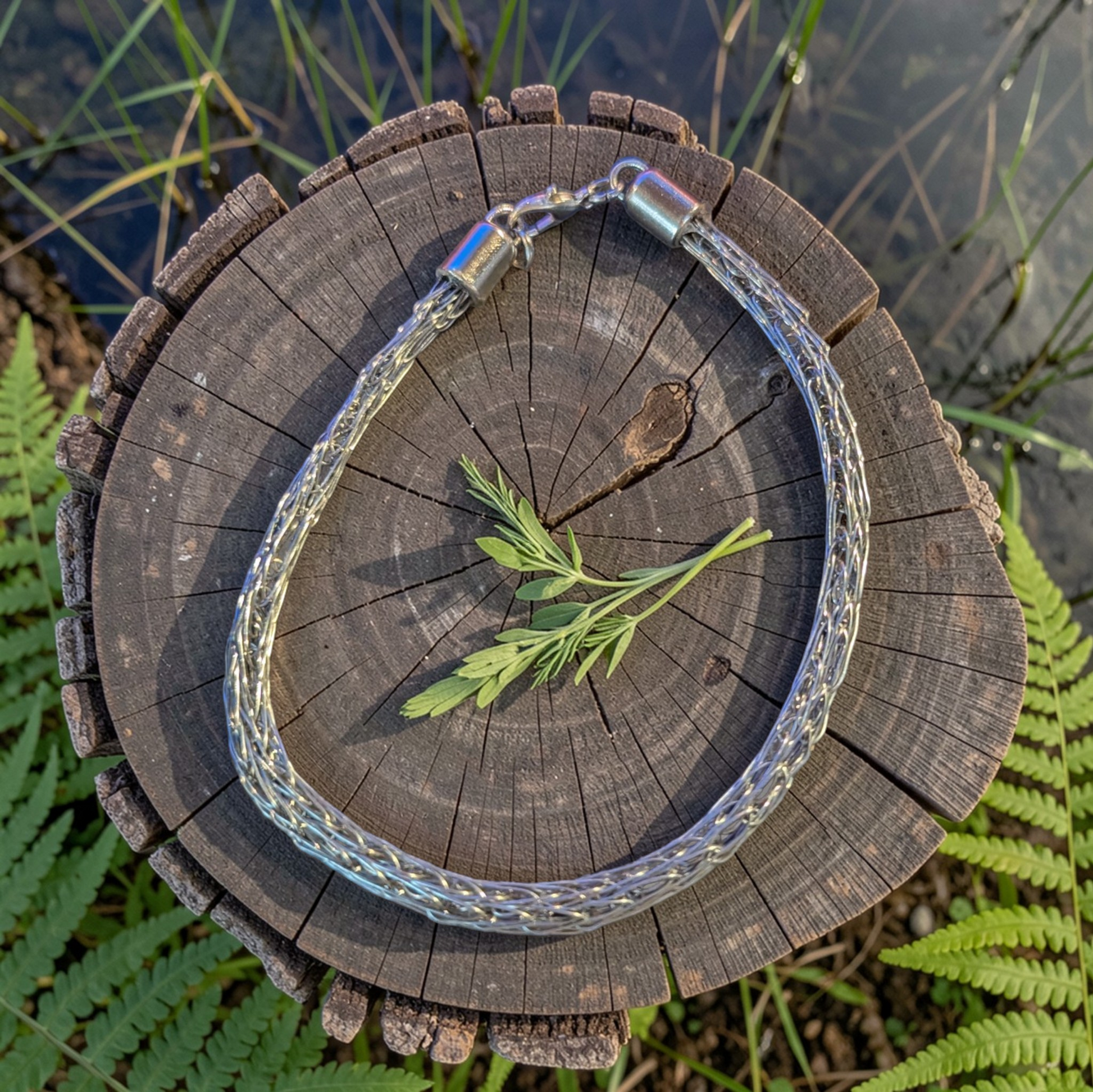 Stainless Steel Trichinopoly weave bracelet arranged in a circle on top of a weathered tree stump outdoors. The bracelet features a dense braided metal weave and a silver-toned lobster claw  clasp at the top. A small green plant sprig rests in the center of the bracelet, with water, grass, and ferns visible around the stump in the background.