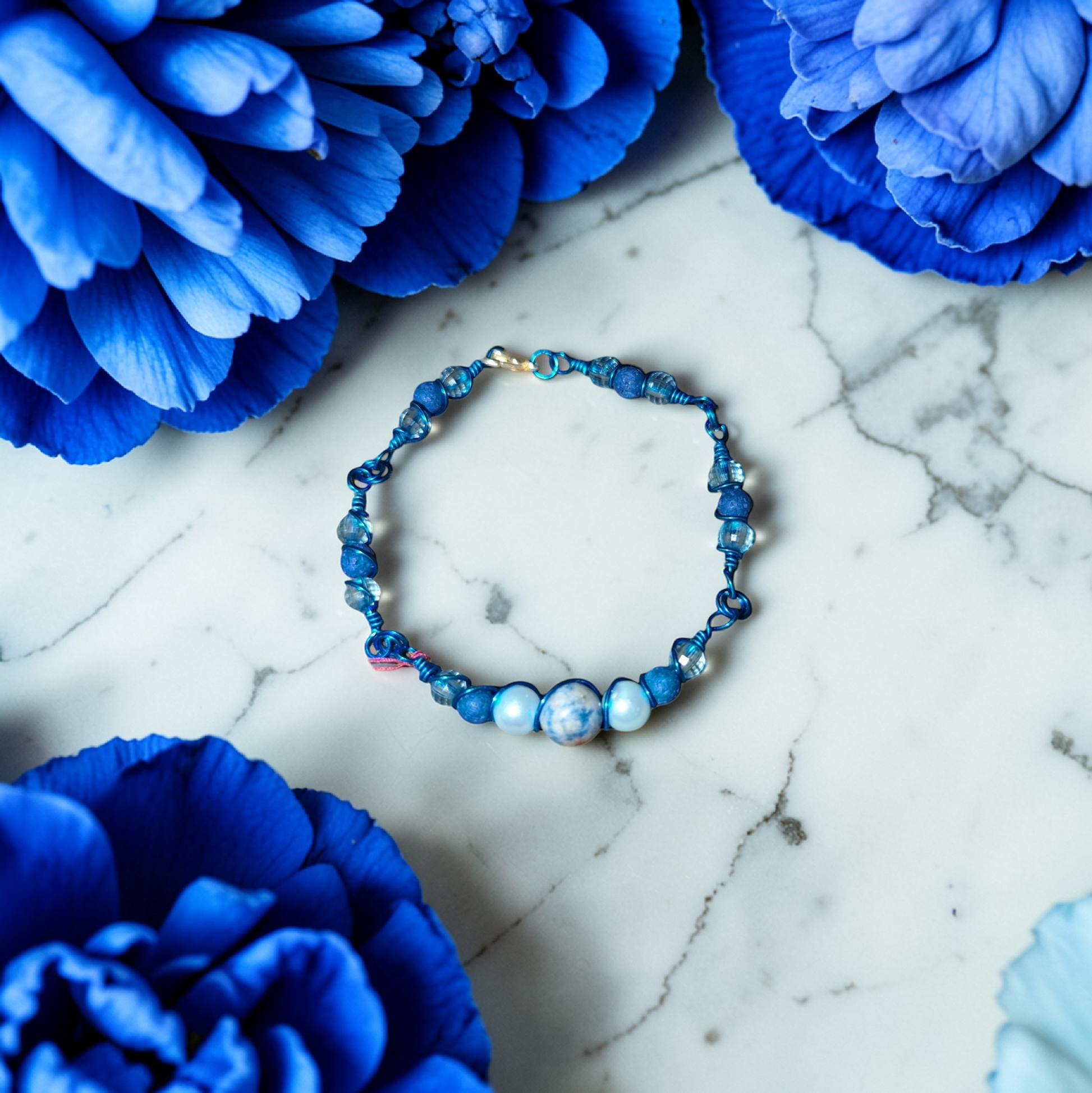 A blue wire-linked bracelet lies centered on a white marble surface, surrounded by large deep-blue flowers framing the edges of the image. The bracelet features a round marbled blue-and-white Sodalite center bead flanked by pale blue pearl-like beads and darker blue lava beads and clear rondelle bead accents, all held in place by twisted blue wire links in a series of links.