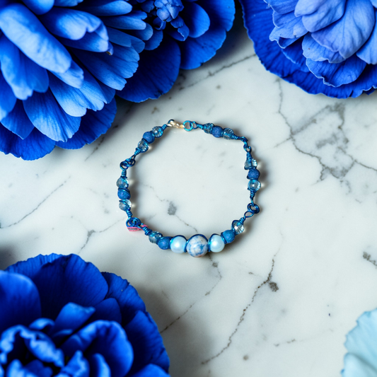 A blue wire-linked bracelet lies centered on a white marble surface, surrounded by large deep-blue flowers framing the edges of the image. The bracelet features a round marbled blue-and-white Sodalite center bead flanked by pale blue pearl-like beads and darker blue lava beads and clear rondelle bead accents, all held in place by twisted blue wire links in a series of links.