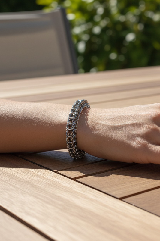 forearm resting on a wooden slat table outdoors: wrist wearing a dense chainmail bracelet forming a thick flexible band; polished gunmetal finish with reflective highlights; tightly woven pattern with repeating V-shaped links wrapping the wrist; toggle clasp partially visible underneath; bright natural sunlight casting sharp shadows; blurred greenery and patio chair in background