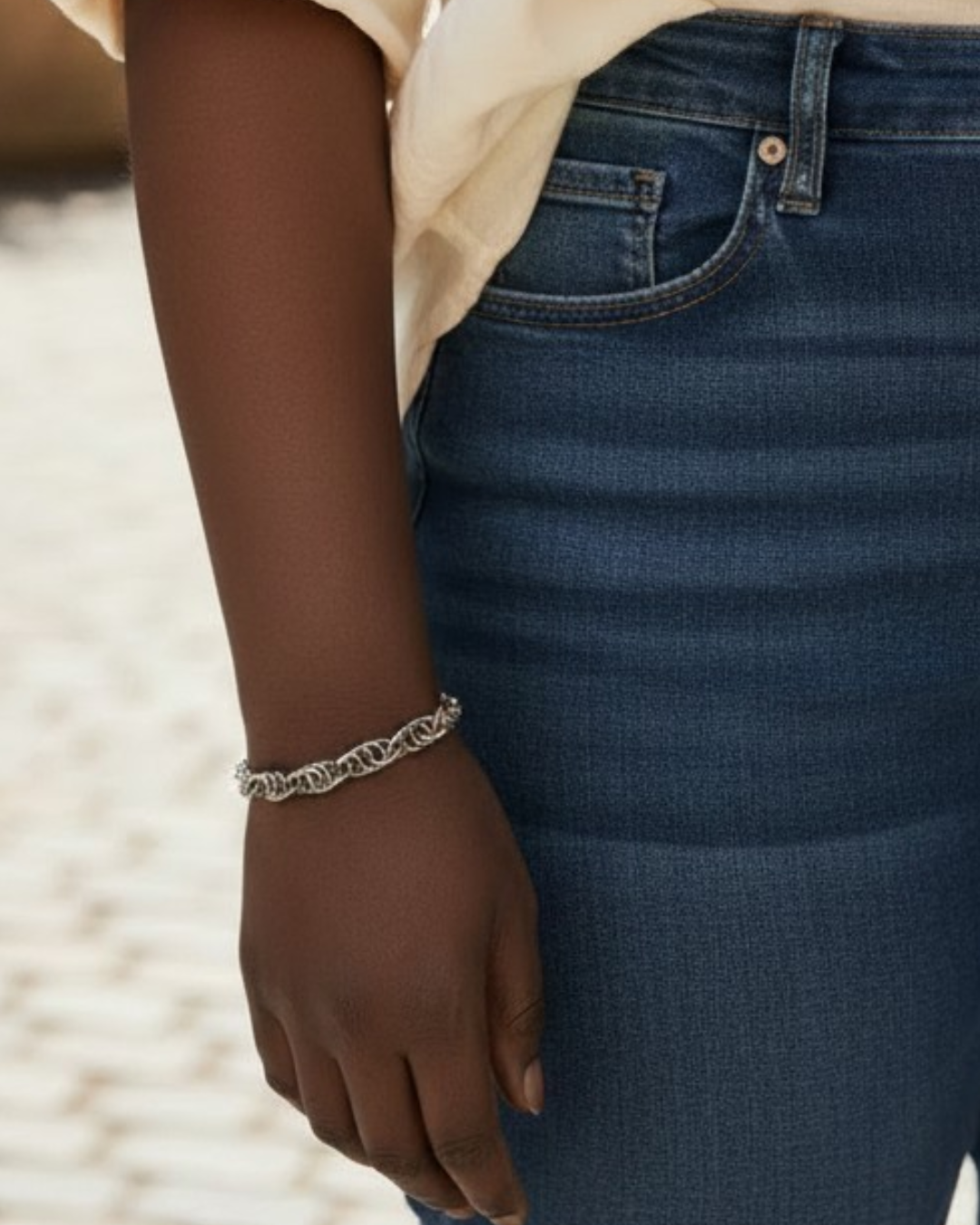 Virtual try-on for scale showing Vinda, a spiral chainmail bracelet with interlocked gun-metal black rings in a hematite-like finish, worn on the wrist of a dark skin tone model. The close-up image shows the bracelet against the wrist beside blue jeans and a cream shirt, with bright outdoor light and a softly blurred background.