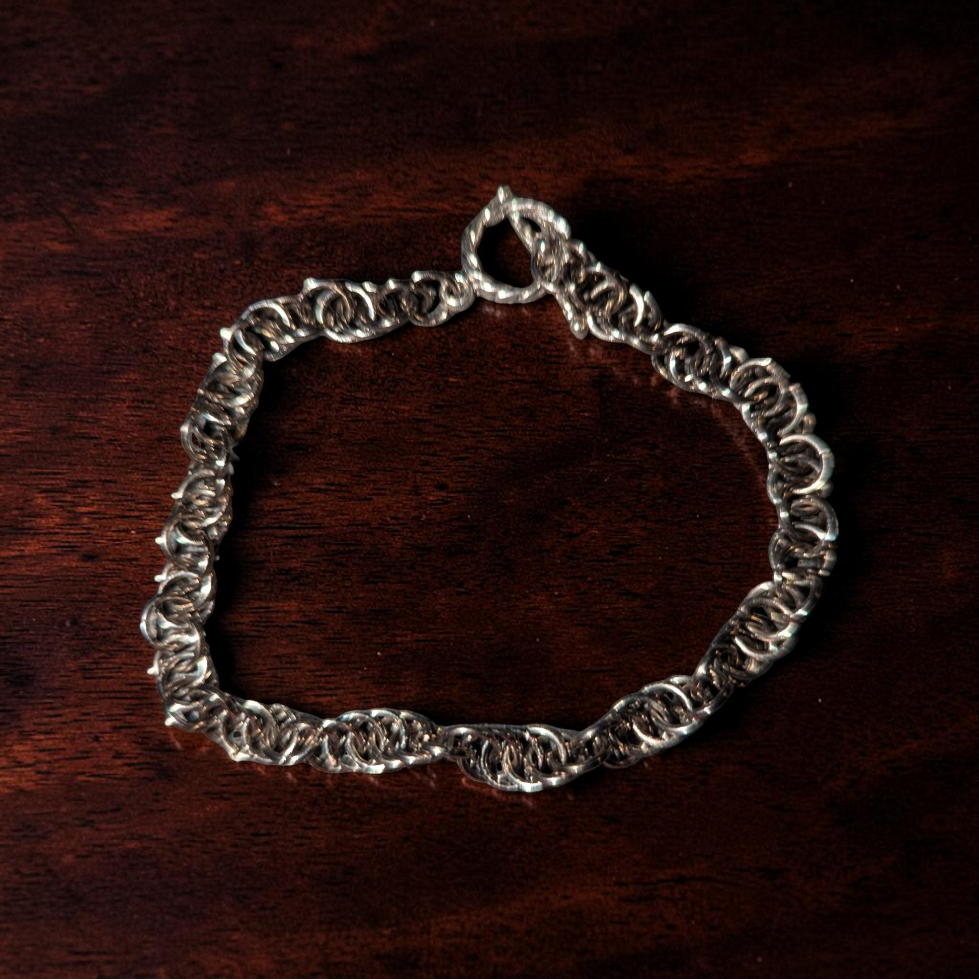 Close-up of Vinda, a spiral chainmail bracelet arranged in an oval on dark reddish-brown wood. The bracelet has interlocked gun-metal black rings with a hematite-like finish, forming a twisted spiral chainmail pattern. A small clasp and connector rings are visible at the top, with shadows emphasizing the bracelet’s raised texture.