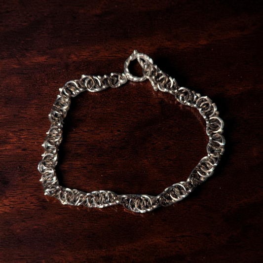 Close-up of Vinda, a spiral chainmail bracelet arranged in an oval on dark reddish-brown wood. The bracelet has interlocked gun-metal black rings with a hematite-like finish, forming a twisted spiral chainmail pattern. A small clasp and connector rings are visible at the top, with shadows emphasizing the bracelet’s raised texture.