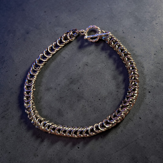 chainmail bracelet resting flat in a loose circle: tightly interlinked silver-toned rings forming a dense Full Persian 6-in-1 weave; toggle clasp at the top with a twisted ring and straight bar slightly angled; metal catching soft light with subtle highlights and shadows; set on a dark, slightly textured concrete surface with faint speckling