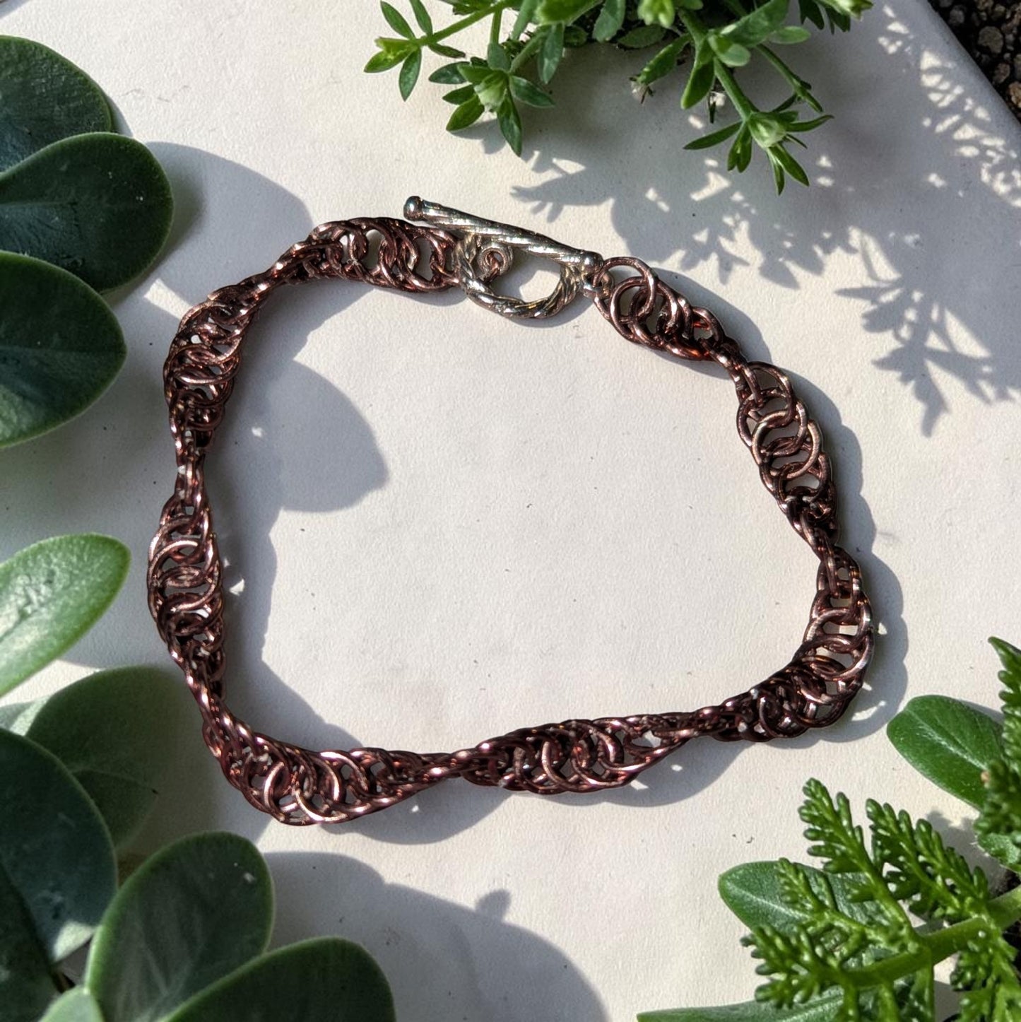 The Snerill spiral 4-in-1 chainmail bracelet in Antique-Bronze laid in a loop on a pale stone surface, surrounded by succulent leaves and ferns, its bronze-toned aluminum rings twisting in a continuous helix and finished with a silver toggle clasp.