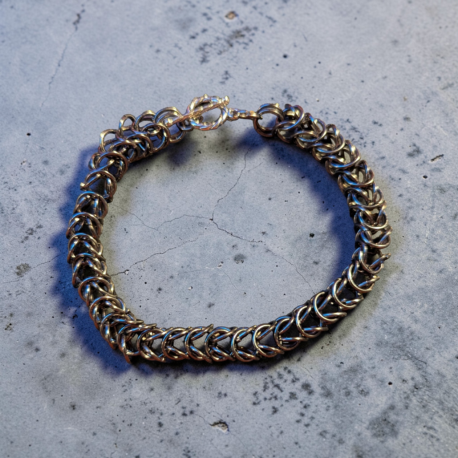 circular bracelet resting on a cracked gray concrete surface: dense interlinked chainmail rings forming a thick flexible band; polished gunmetal finish with reflective highlights; tightly woven pattern with repeating V-shaped links; toggle clasp at top with metal loop and straight bar; soft directional light casting faint shadows into the weave and across the concrete texture
