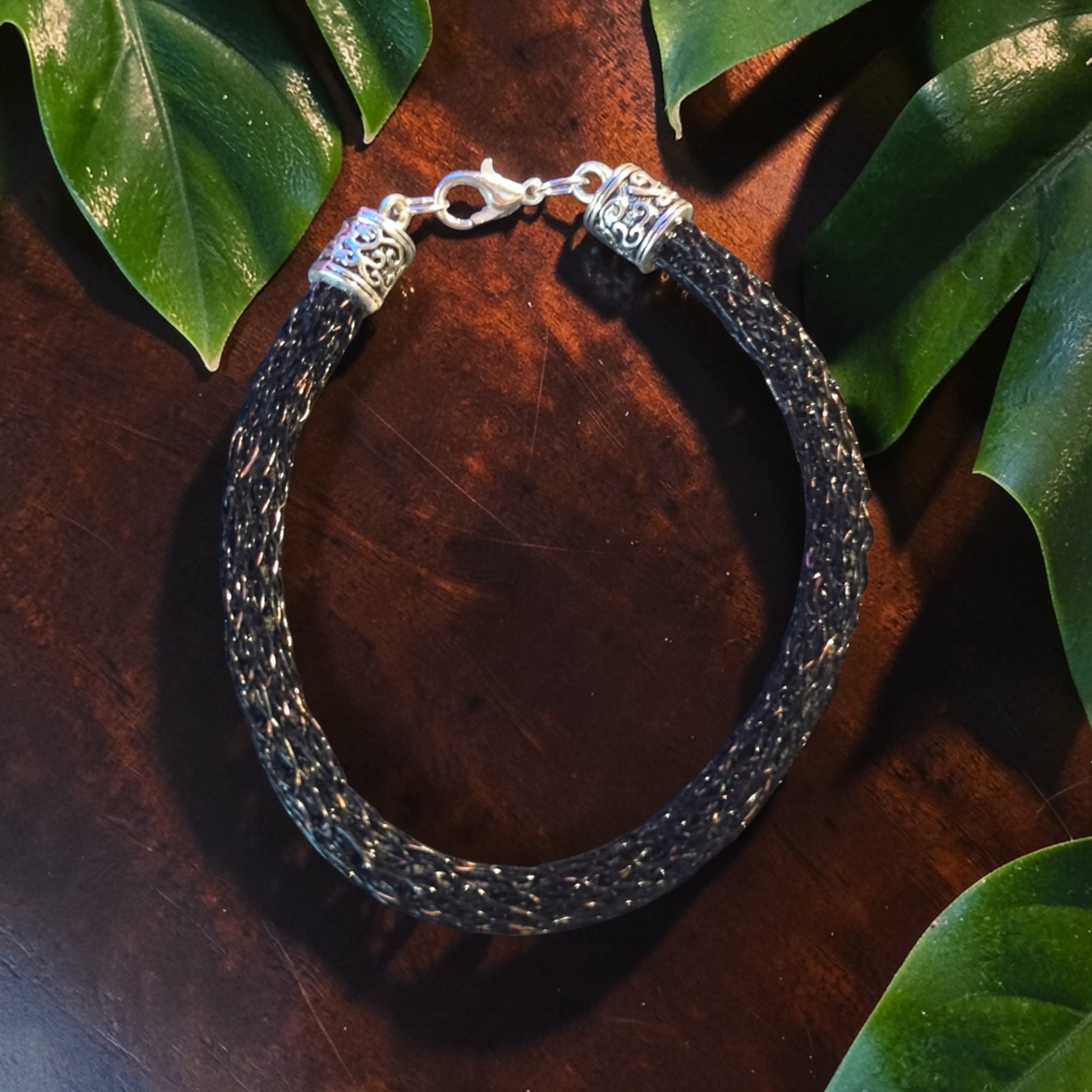 The image shows a Viking knit bracelet made from distressed black lacquered copper wire forming a dense woven tube. The bracelet rests in a circular shape on a dark wooden surface surrounded by green leaves. Each end is finished with antique silver Tibetan-style end caps connected by a silver lobster clasp. The textured woven wire catches light subtly against the darker finish, highlighting the knit pattern and metal fittings.