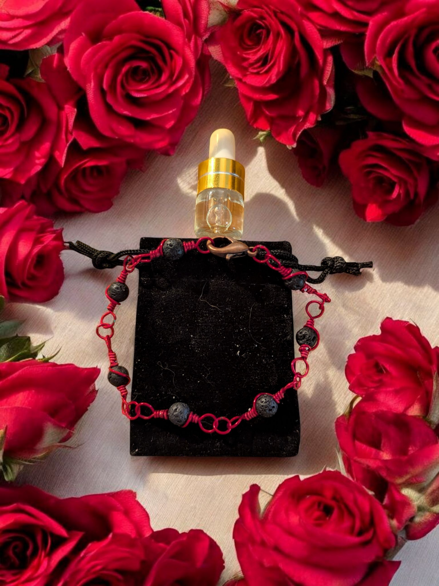 Handcrafted black lava diffuser bracelet with red hypoallergenic wire, displayed on a black velvet pouch surrounded by red roses and a small aromatherapy vial.