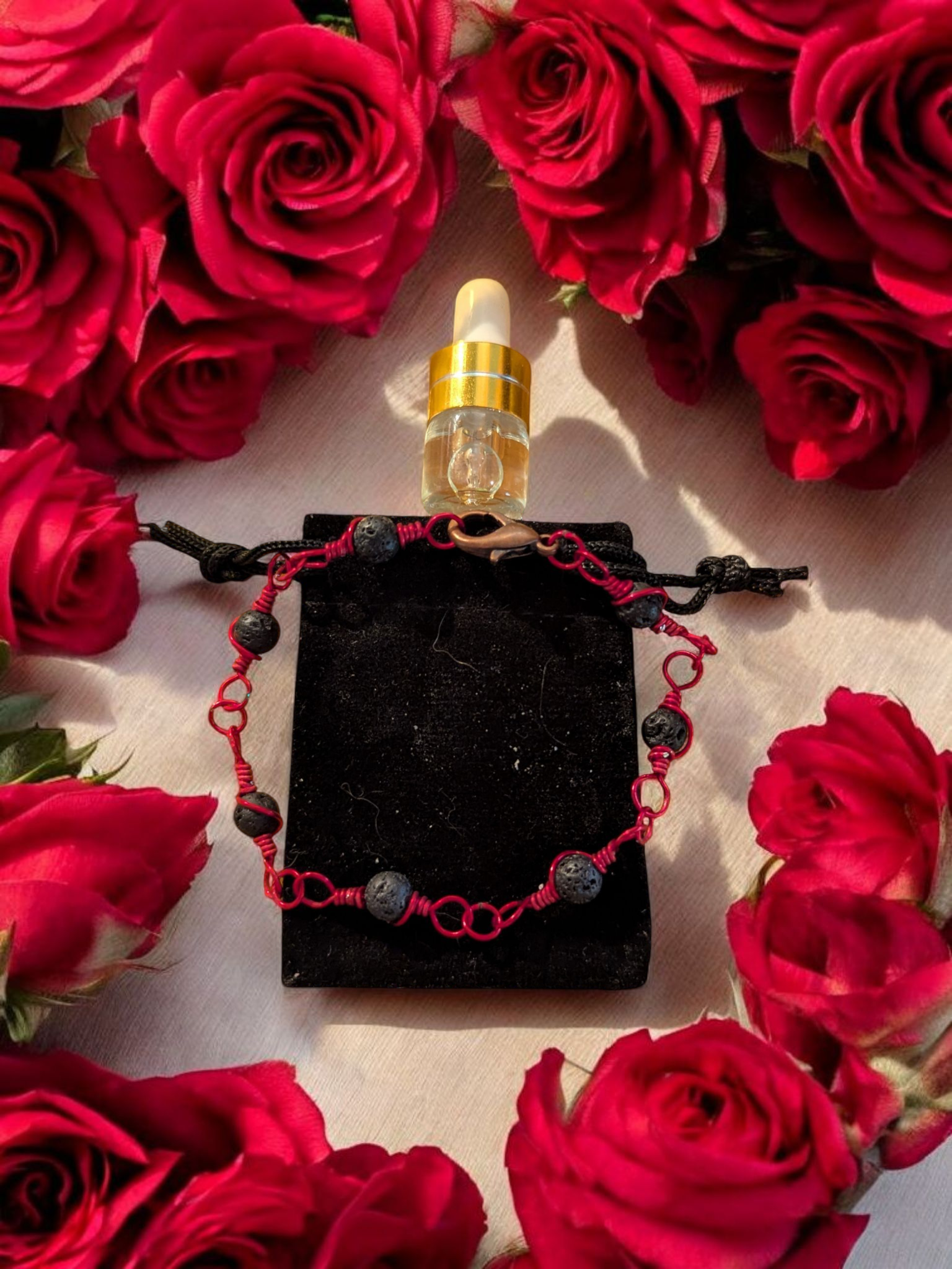 Handcrafted black lava diffuser bracelet with red hypoallergenic wire, displayed on a black velvet pouch surrounded by red roses and a small aromatherapy vial.